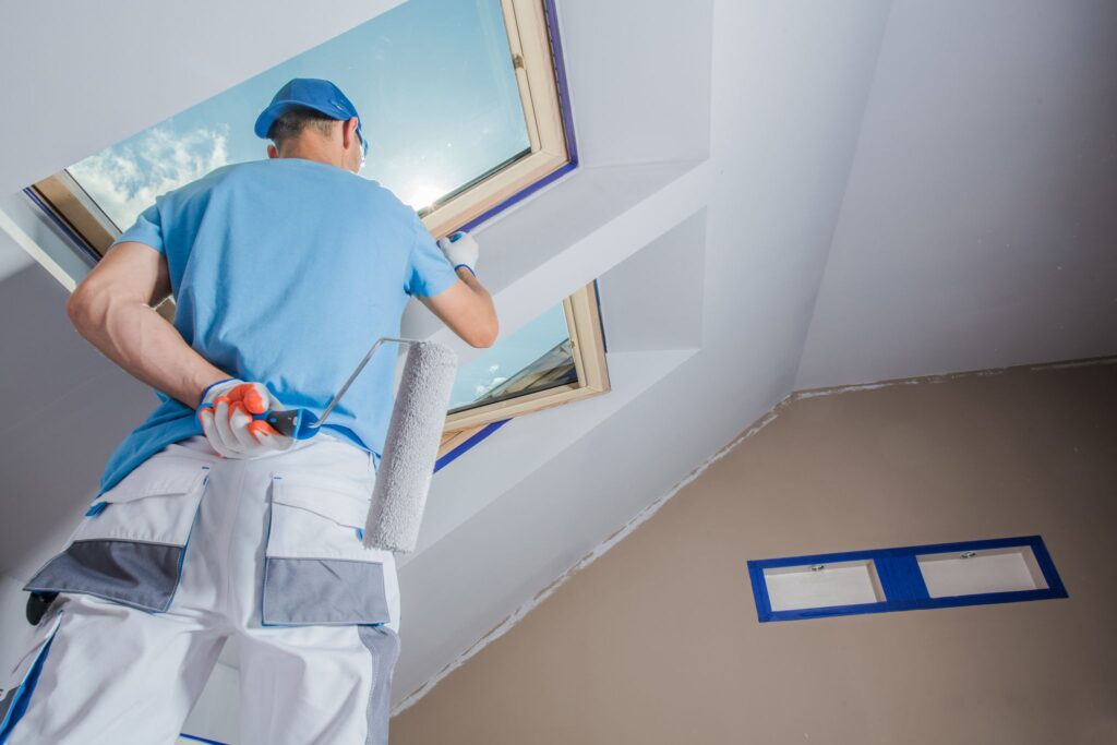 A man painting a room with a skylight.